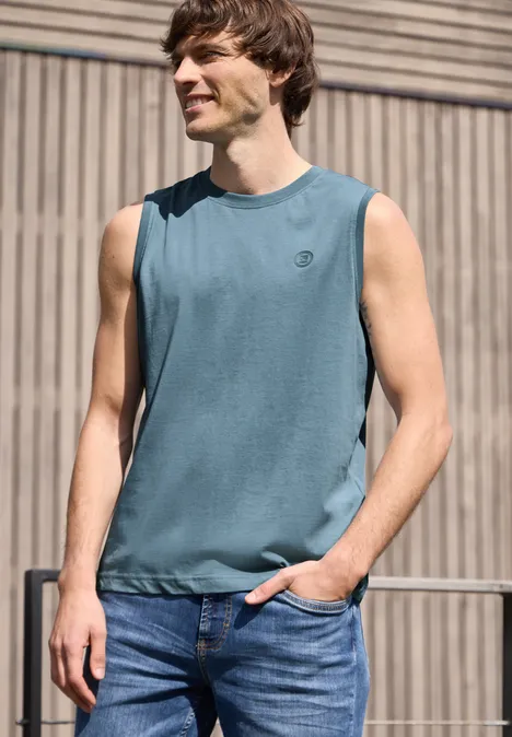 Een man in een mouwloos T-shirt en jeans staat ontspannen voor een houten gevel.