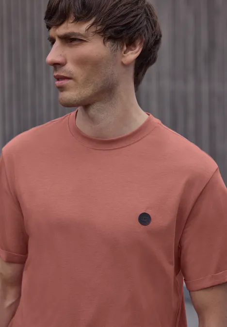 Ein verschwommenes Porträt einer Person in rostfarbenem T-Shirt vor einer grauen, vertikal gestreiften Wand.