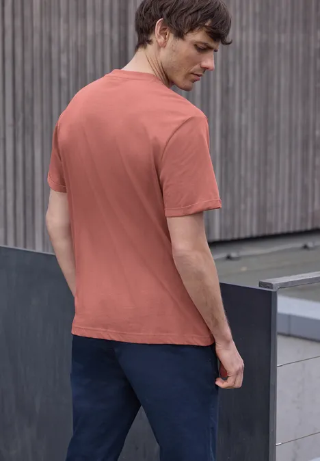 Een man van achteren in een baksteenrood t-shirt en donkere broek voor een houten gevel.