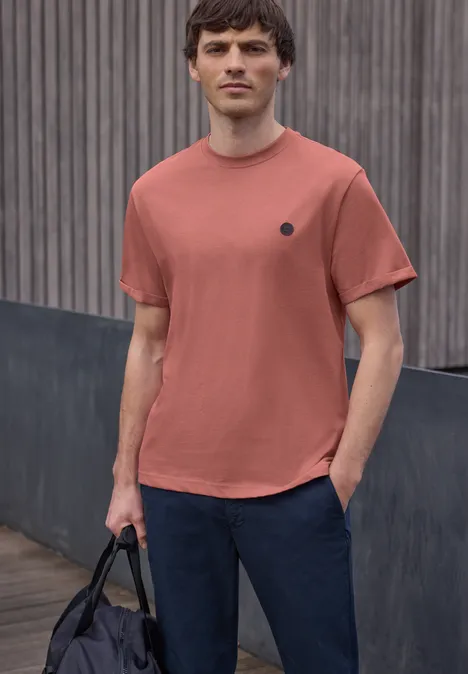 Een man draagt een roestkleurig t-shirt en donkere broek, met een zwarte tas voor een houten wand.