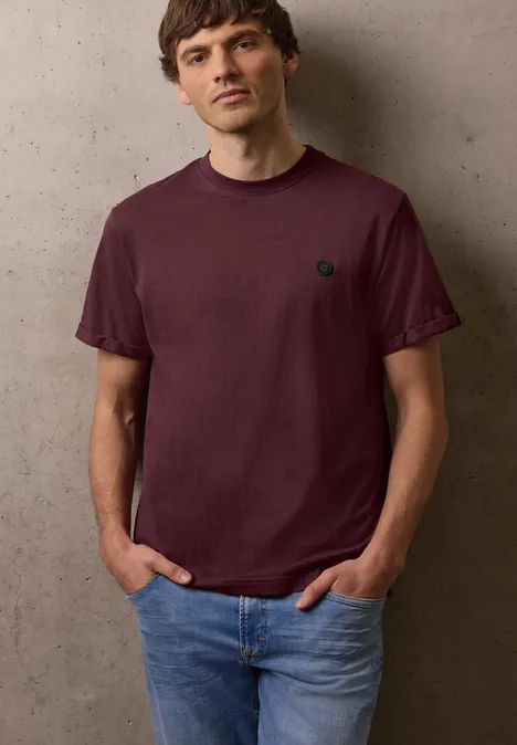 Mann lehnt an grauer Wand, trägt bordeauxrotes T-Shirt mit kleinem Logo und blauen Jeans, Hände in den Taschen.