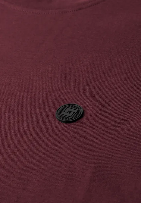 Een bordeauxrood T-shirt met een klein, zwart rond embleem op de borst.