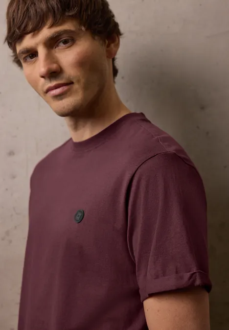 Een man draagt een eenvoudig paars T-shirt met opgerolde mouwen en een klein rond logo op de borst.