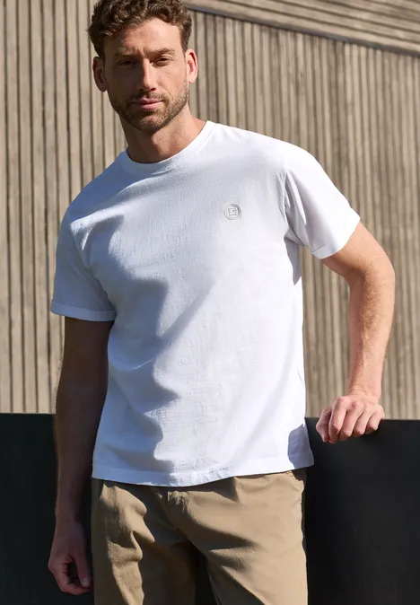 Ein Mann in weißem T-Shirt und beigen Hosen steht vor einer Holzfassade.