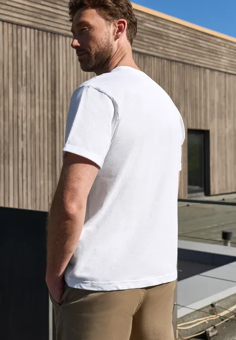 Ein Mann steht rückwärts gekehrt auf einer Terrasse, trägt ein weißes T-Shirt und khakifarbene Hose vor Holzfassade.