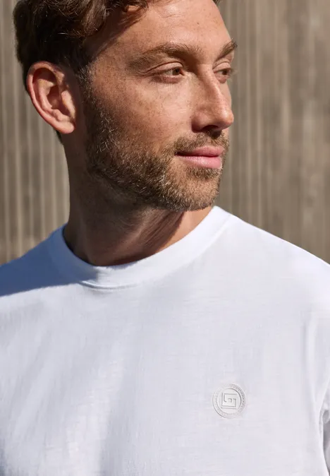 Ein Mann in weißem T-Shirt mit kleinem Logo steht vor einer Holzwand