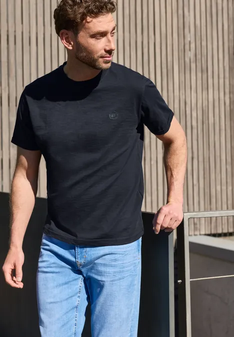 Ein Mann steht lässig in einem schwarzen T-Shirt und blauen Jeans vor einer Holzwand.