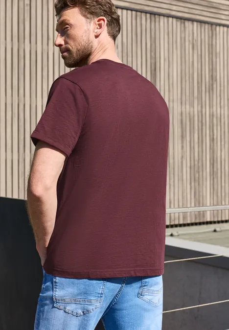 Ein Mann in bordeauxrotem T-Shirt und blauen Jeans steht seitlich mit dem Rücken zur Kamera vor einer Holzwand.