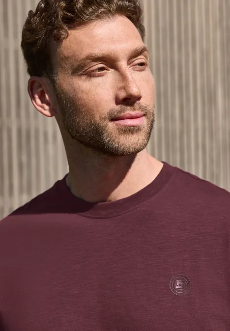 Ein Mann mit verschwommenem Gesicht trägt ein dunkelrotes T-Shirt vor einer Holzlattenwand.