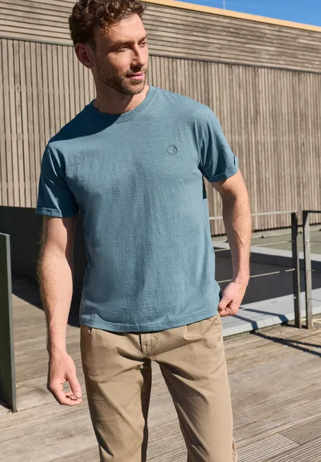 Ein Mann in blauem T-Shirt und beigen Hosen steht auf einer Holzterrasse vor einer Holzfassade.