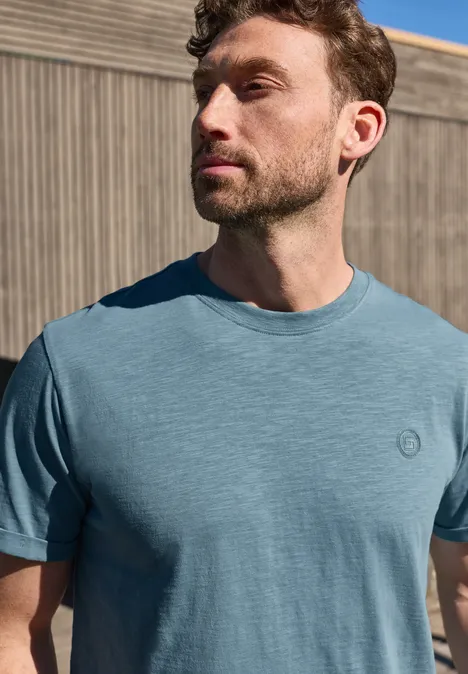 Mann steht vor Holzwand in blauem T-Shirt, schaut seitlich.