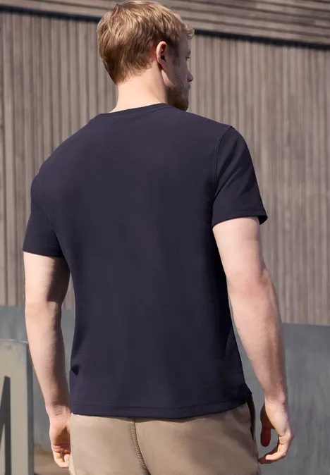 Een man met zijn rug naar de camera, gekleed in een donkerblauw T-shirt en beige broek, staand voor een houten wand.