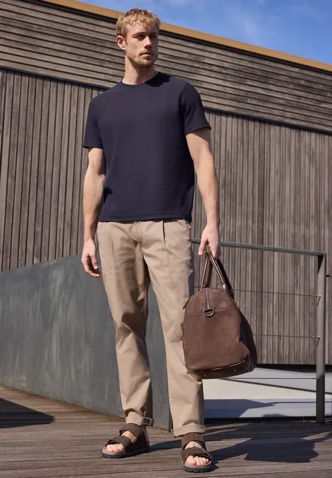 Een man voor een houten gevel in een zwart T-shirt, beige broek en sandalen, met een bruine tas in zijn hand.