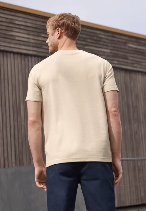 Een man staat met zijn rug naar de camera en draagt een beige t-shirt en een donkere broek voor een houten wand.