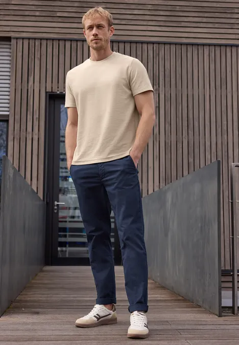 Een man staat voor een houten wand, draagt een beige t-shirt, blauwe broek en witte sneakers.
