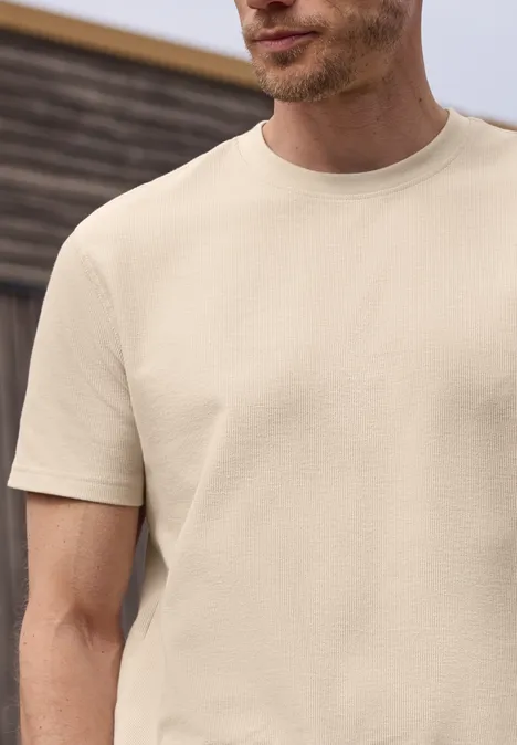 Een man draagt een beige t-shirt met korte mouwen tegen een onscherpe houten achtergrond.