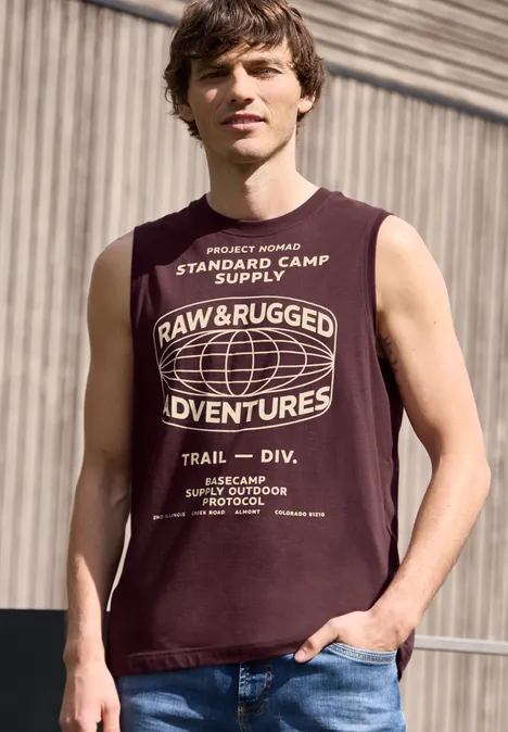 Ein Mann in ärmellosem, dunkelrotem T-Shirt mit weißem Aufdruck 'Raw & Rugged Adventures' vor Industriehintergrund.