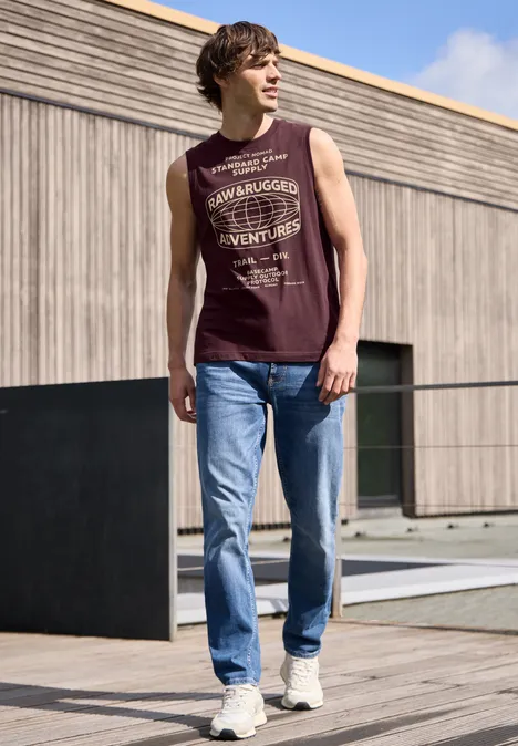 Een jonge man in mouwloos shirt en spijkerbroek loopt over een houten terras voor een modern huis.