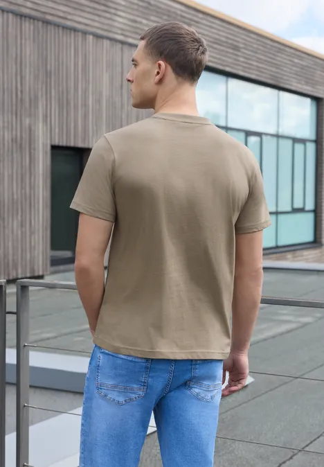 Ein Mann in Beige-T-Shirt und Jeans steht auf einer Dachterrasse, schaut auf moderne Glasfassade.