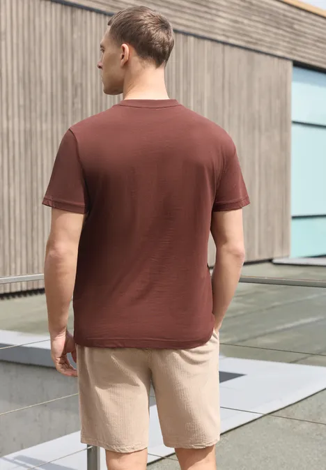 Ein Mann mit braunem T-Shirt und beigen Shorts steht mit dem Rücken zur Kamera vor einem modernen Gebäude.
