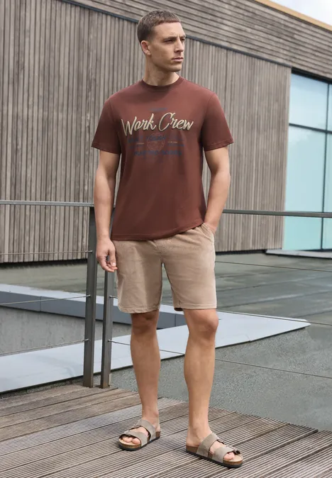Mann in braunem T-Shirt und beigen Shorts steht auf Holzterrasse vor modernem Gebäude.