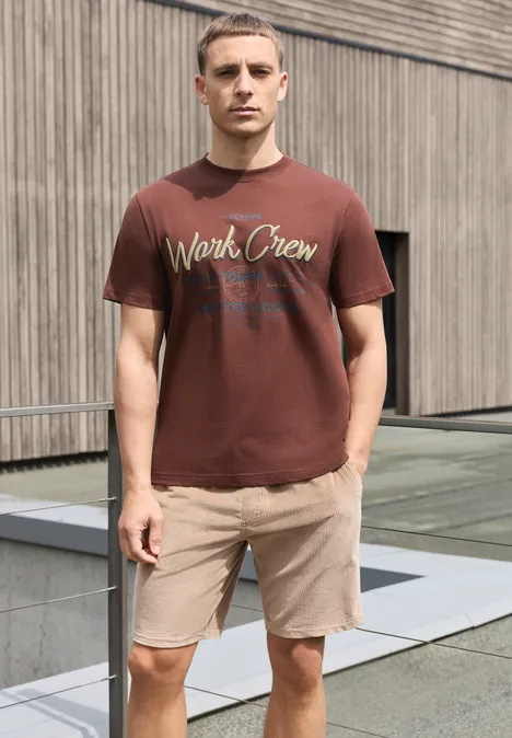 Ein Mann in braunem T-Shirt und beigen Shorts steht vor moderner Holzwand.