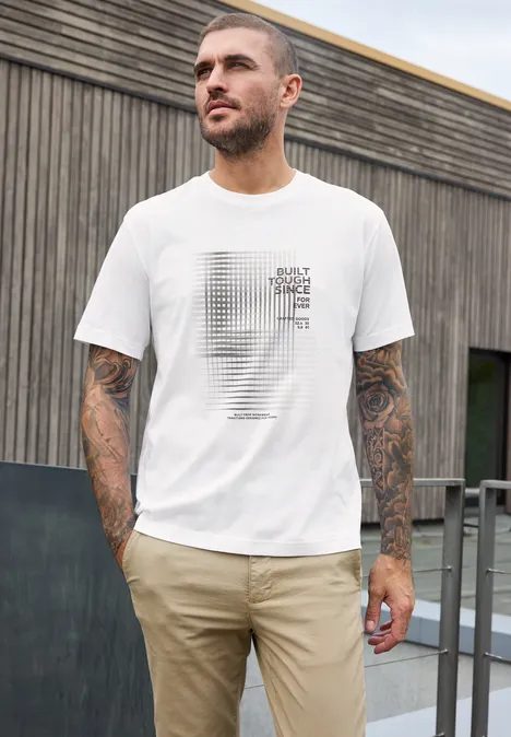 Ein tätowierter Mann mit weißem T-Shirt und beigen Hosen steht draußen vor einer Holzwand
