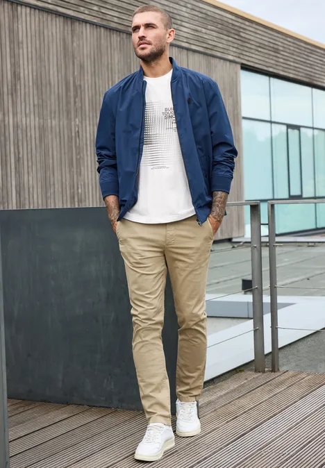 Ein Mann in beige Chinos, weißem T-Shirt und blauer Bomberjacke geht auf einer Holzterrasse.