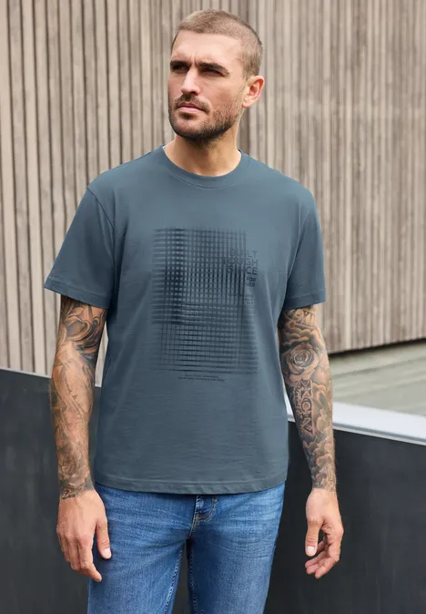 Ein tätowierter Mann in T-Shirt und Jeans steht vor einer Holzwand und blickt zur Seite.