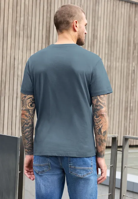 Ein Mann mit Tattoos auf beiden Armen, trägt graues T-Shirt und Jeans, steht Rücken zur Kamera vor einer Holzfassade.