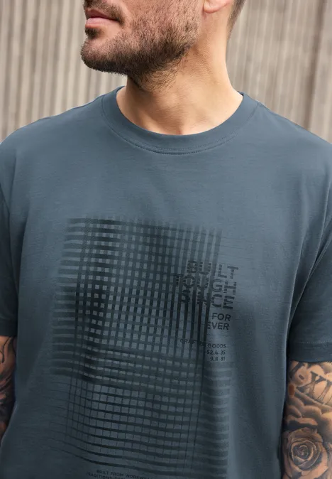 Ein Mann mit Bart trägt ein graublaues T-Shirt mit Rastermuster und Aufdruck, sichtbares Rosen-Tattoo am Arm.