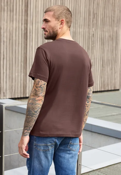 Ein tätowierter Mann im braunen T-Shirt und Jeans steht vor einer Holzfassade im Freien.