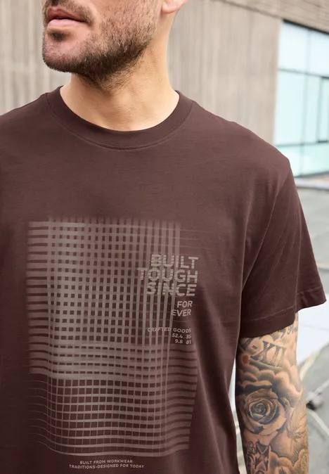 Mann trägt braunes T-Shirt mit Gitter-Design und Schriftzug, sichtbarer tätowierter Unterarm