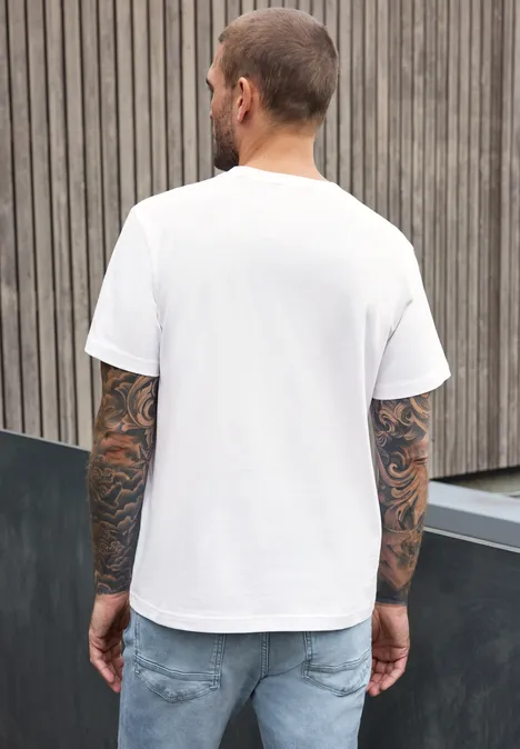 Ein tätowierter Mann in weißem T-Shirt und Jeans, Rückenansicht vor Holzwand.