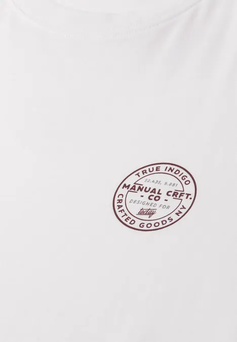 Ein weißes T-Shirt mit ovalem roten Logo-Aufdruck "Manual Crafted Co Limited Goods" auf Brusthöhe.