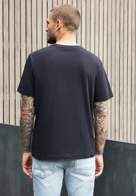 Ein Mann mit Tattoos trägt ein schwarzes T-Shirt und Jeans, steht vor einer Holzlattenwand, Rückenansicht.