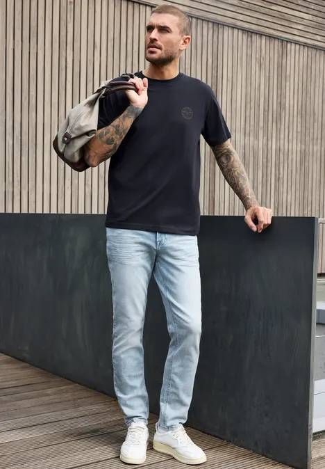 Ein Mann in schwarzem T-Shirt, hellblauen Jeans und weißen Sneakern trägt eine Tasche über der Schulter vor einer Holzfassade.