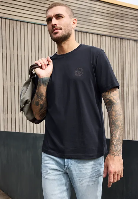Ein tätowierter Mann trägt ein schwarzes T-Shirt und hellblaue Jeans, hält lässig einen Rucksack über der Schulter.