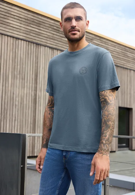 Ein Mann in blauem T-Shirt und Jeans mit Tattoos an den Armen steht vor einem modernen Holzgebäude.