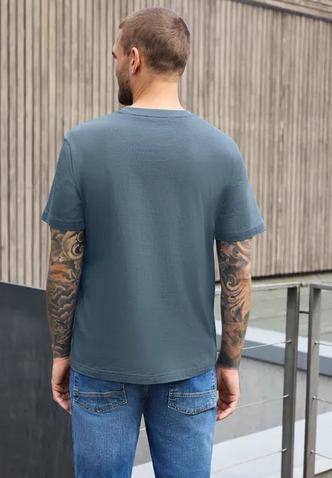 Ein tätowierter Mann von hinten in blauem T-Shirt und Jeans lehnt an einem Holzzaun.