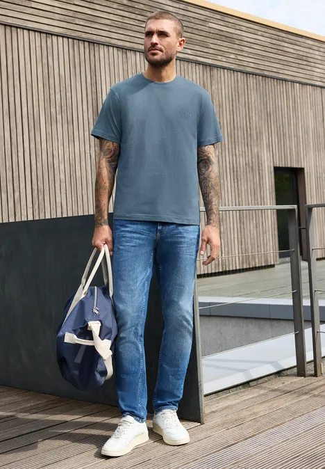 Ein Mann auf einem Holzdeck trägt blaues T-Shirt, Jeans, weiße Sneaker und hält eine blaue Reisetasche.
