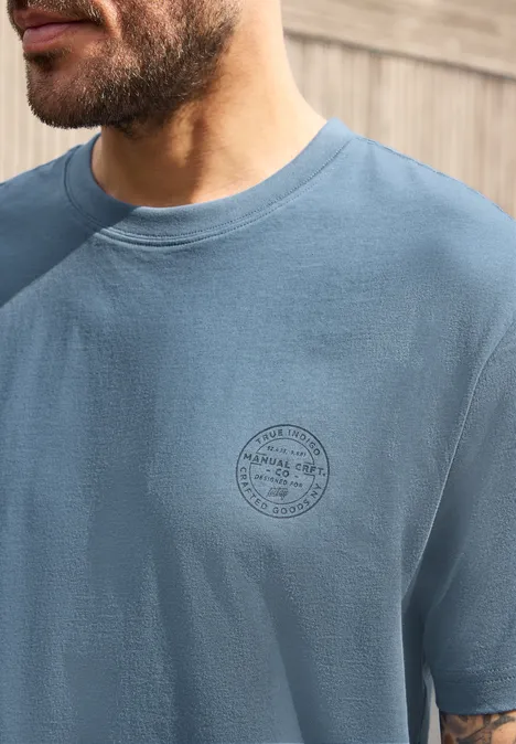 Nahaufnahme eines bärtigen Manns in blauem T-Shirt mit rundem Logo auf der Brust vor Holzverkleidung.