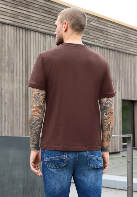 Ein Mann steht mit dem Rücken zur Kamera, trägt braunes T-Shirt und Jeans vor modernem Holzgebäude.