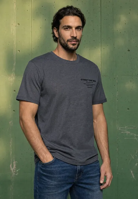 Ein Mann mit dunklem Haar und Bart trägt ein graues T-Shirt und Jeans vor einem grünen Hintergrund.