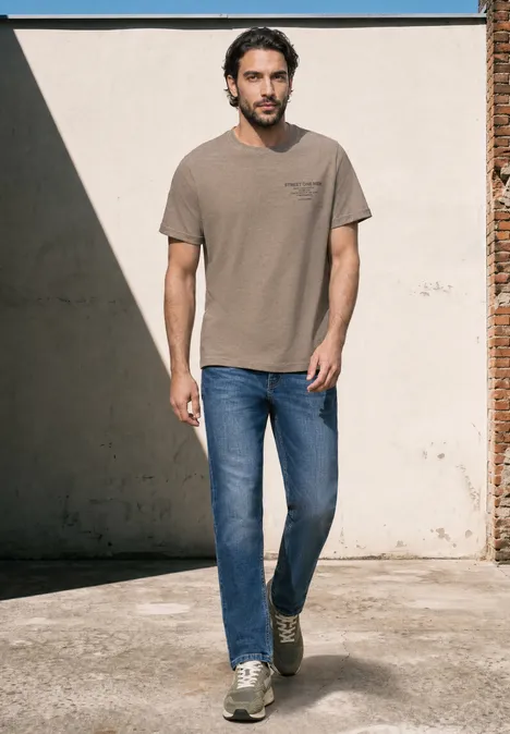 Ein Mann in lässigem T-Shirt, Jeans und Sneakers läuft an einer sonnigen Mauer entlang.