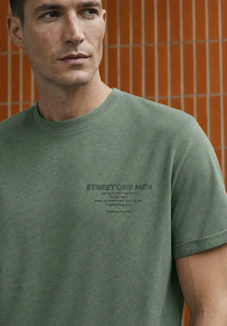 Mann trägt grünes T-Shirt mit kleinem Logo, steht vor orangefarbener Fliesenwand.