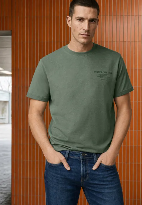 Ein Mann in einem grünen T-Shirt und Jeans steht vor einer orange-braunen Wand mit senkrechten Paneelen.