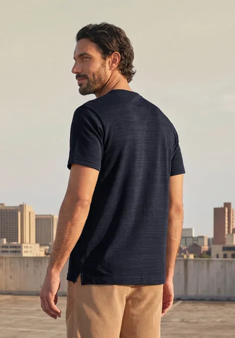 Ein Mann mit dunkelblauer T-Shirt und beige Shorts steht auf einem Dach und blickt über die Stadt.