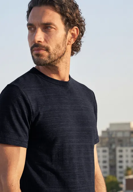 Ein bärtiger Mann mit dunklem T-Shirt blickt seitlich vor urbaner Kulisse.