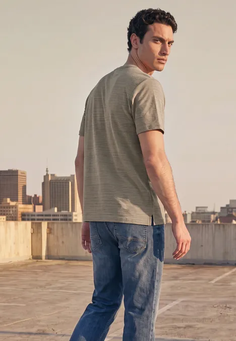 Ein Mann in T-Shirt und Jeans steht auf einem Dach, Blick über die Stadt im Hintergrund.